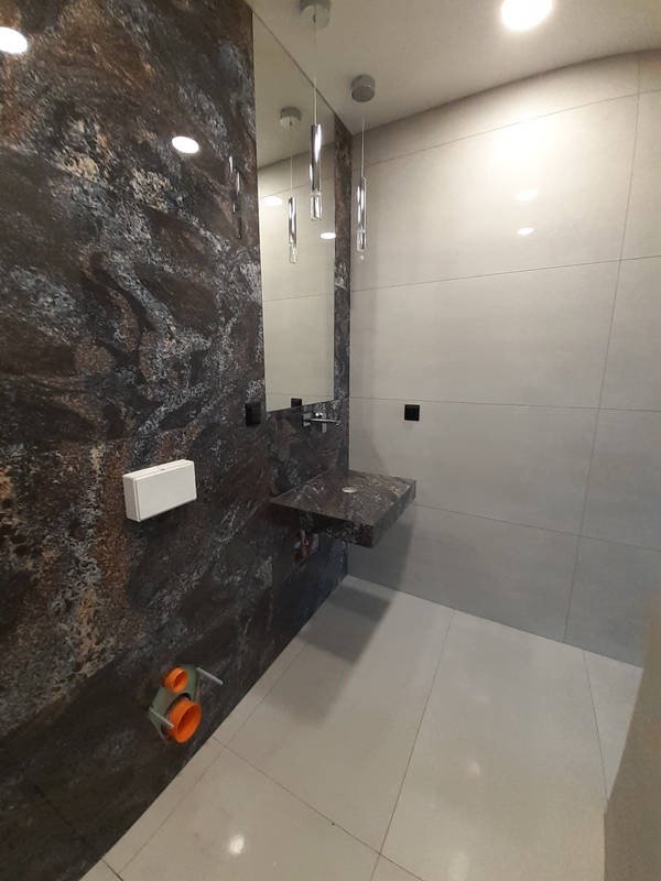 Nowoczesna łazienka z umywalką z ciemnego kamienia, ściana wyłożona płytkami imitującymi marmur i wiszącymi lampami, widoczne elementy instalacji wodnej przed zabudową.