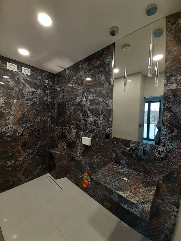 Nowoczesna łazienka z ciemnymi, marmurkowymi płytkami na ścianach i blacie umywalki, jasne płytki podłogowe, minimalistyczne oświetlenie sufitowe i pionowe lampy wiszące odbijające się w lustrze...