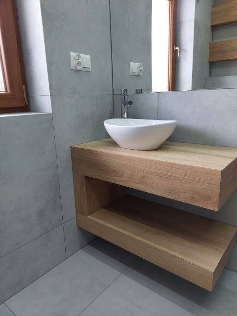 Nowoczesna łazienka z umywalką nablatową w kształcie misy, zamontowaną na drewnianym blacie z półką. Szare, matowe płytki wielkoformatowe na ścianach i podłodze. W tle widoczne okno z drewnianą...