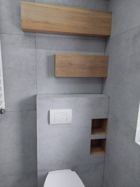 Szara łazienka z podtynkową toaletą, drewnianymi półkami nad nią i dwoma wnękami na ścianie wykończonej płytkami imitującymi beton.