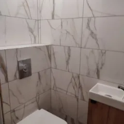 Nowoczesna łazienka z marmurowymi płytkami na ścianach, wiszącą toaletą i umywalką z drewnianą szafką. Widoczny chromowany przycisk spłuczki.