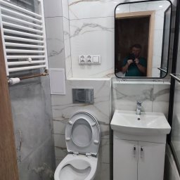 Marek Krasnowski - Nowoczesna łazienka po remoncie: białe marmurowe płytki, wisząca toaleta, szafka z umywalką i lustro z odbiciem fotografa. Grzejnik drabinkowy.