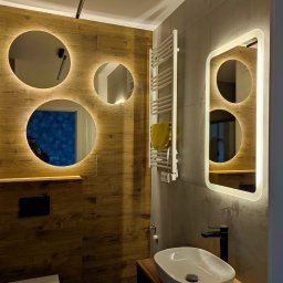 Marek Krasnowski - Nowoczesna łazienka z drewnopodobnymi płytkami, okrągłymi lustrami z podświetleniem LED i owalną umywalką nablatową. Grzejnik drabinkowy na szarej ścianie.