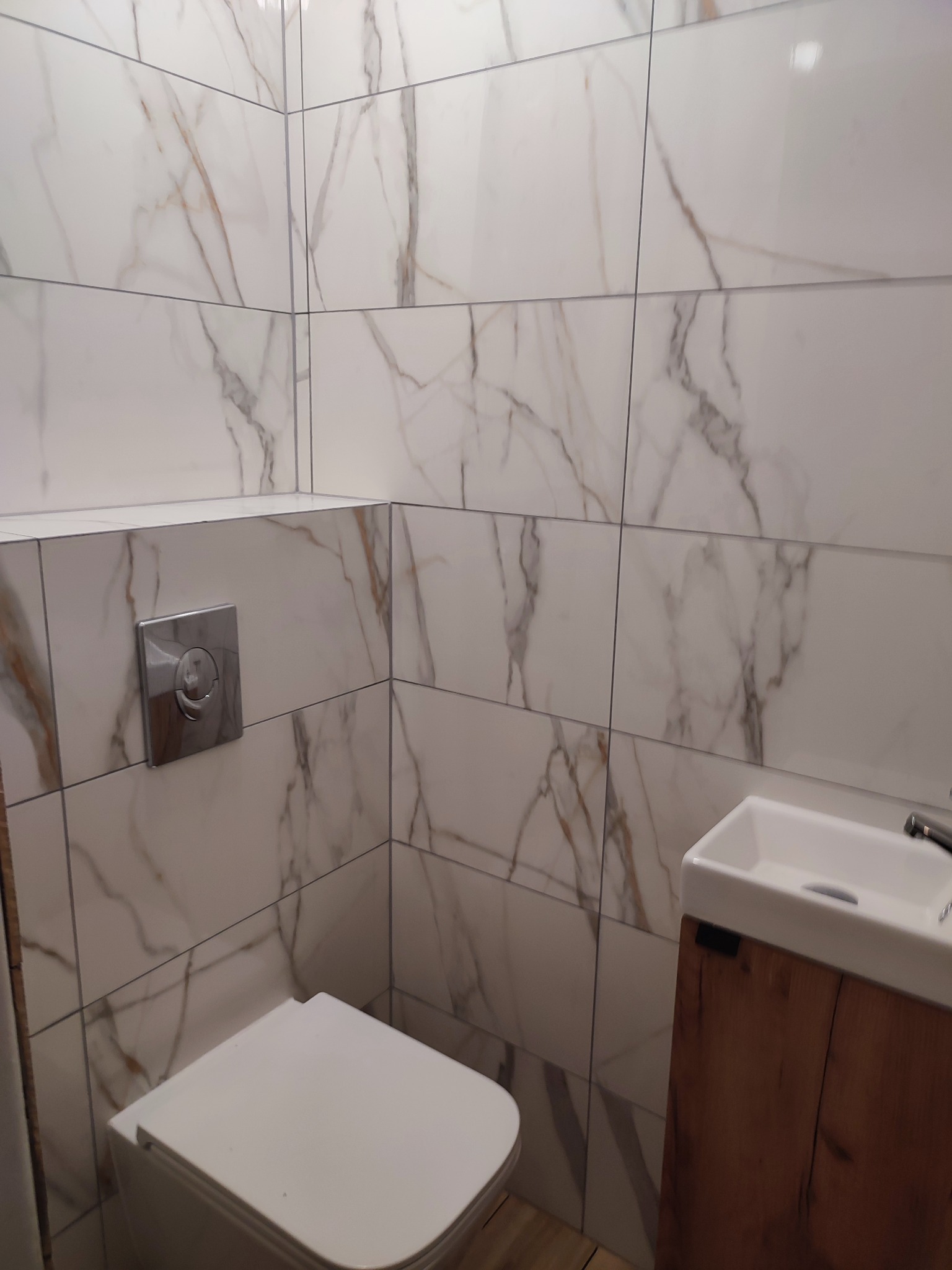 Nowoczesna łazienka z marmurowymi płytkami na ścianach, wiszącą toaletą i umywalką z drewnianą szafką. Widoczny chromowany przycisk spłuczki.