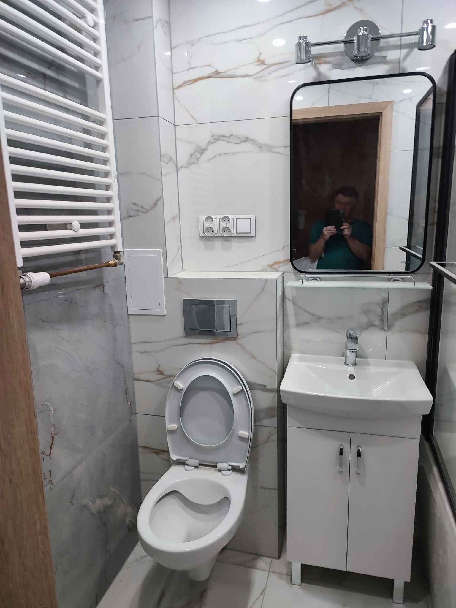 Nowoczesna łazienka po remoncie: białe marmurowe płytki, wisząca toaleta, szafka z umywalką i lustro z odbiciem fotografa. Grzejnik drabinkowy.