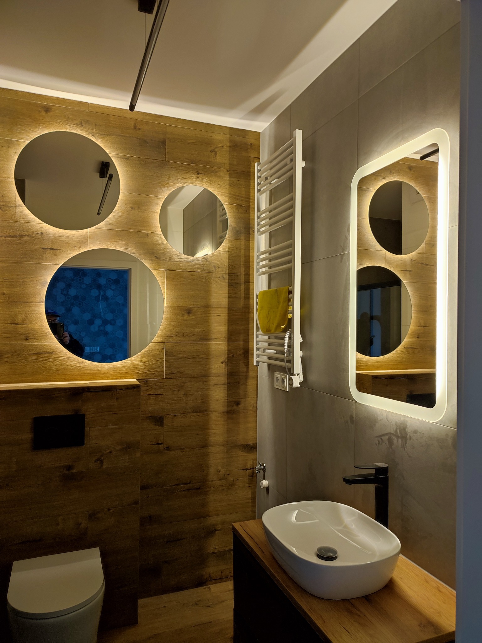 Nowoczesna łazienka z drewnopodobnymi płytkami, okrągłymi lustrami z podświetleniem LED i owalną umywalką nablatową. Grzejnik drabinkowy na szarej ścianie.
