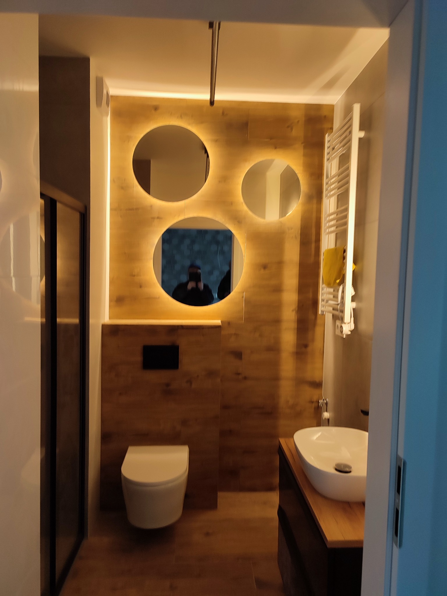 Nowoczesna łazienka w ciepłych odcieniach drewna z okrągłymi lustrami podświetlanymi LED i białą armaturą. Widoczny grzejnik drabinkowy i kabina prysznicowa.