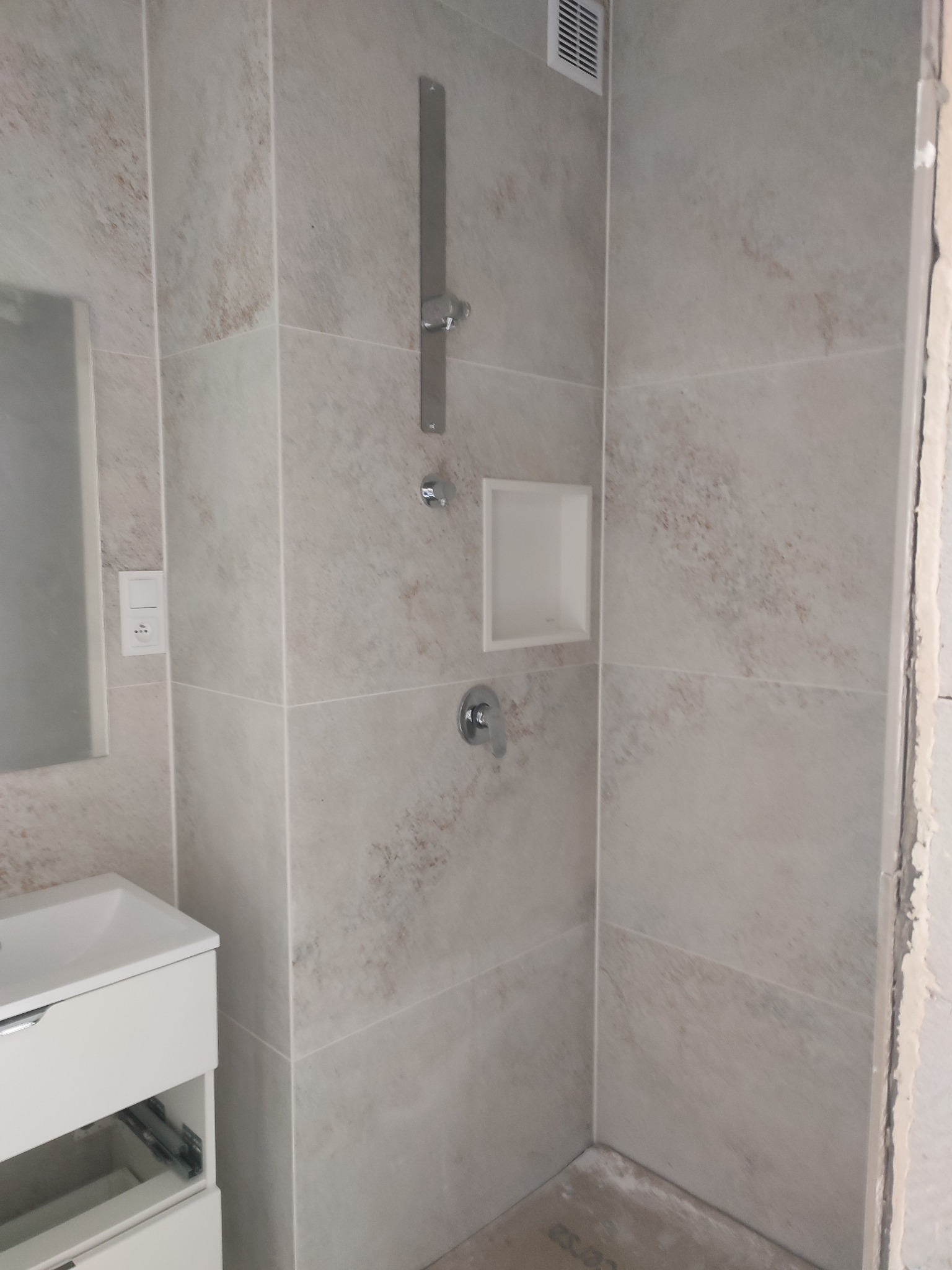 Wykończona kabina prysznicowa w beżowych płytkach z wnęką, nowoczesną armaturą i białą szafką z umywalką. Widoczny wentylator i fragment niezabezpieczonej ściany.