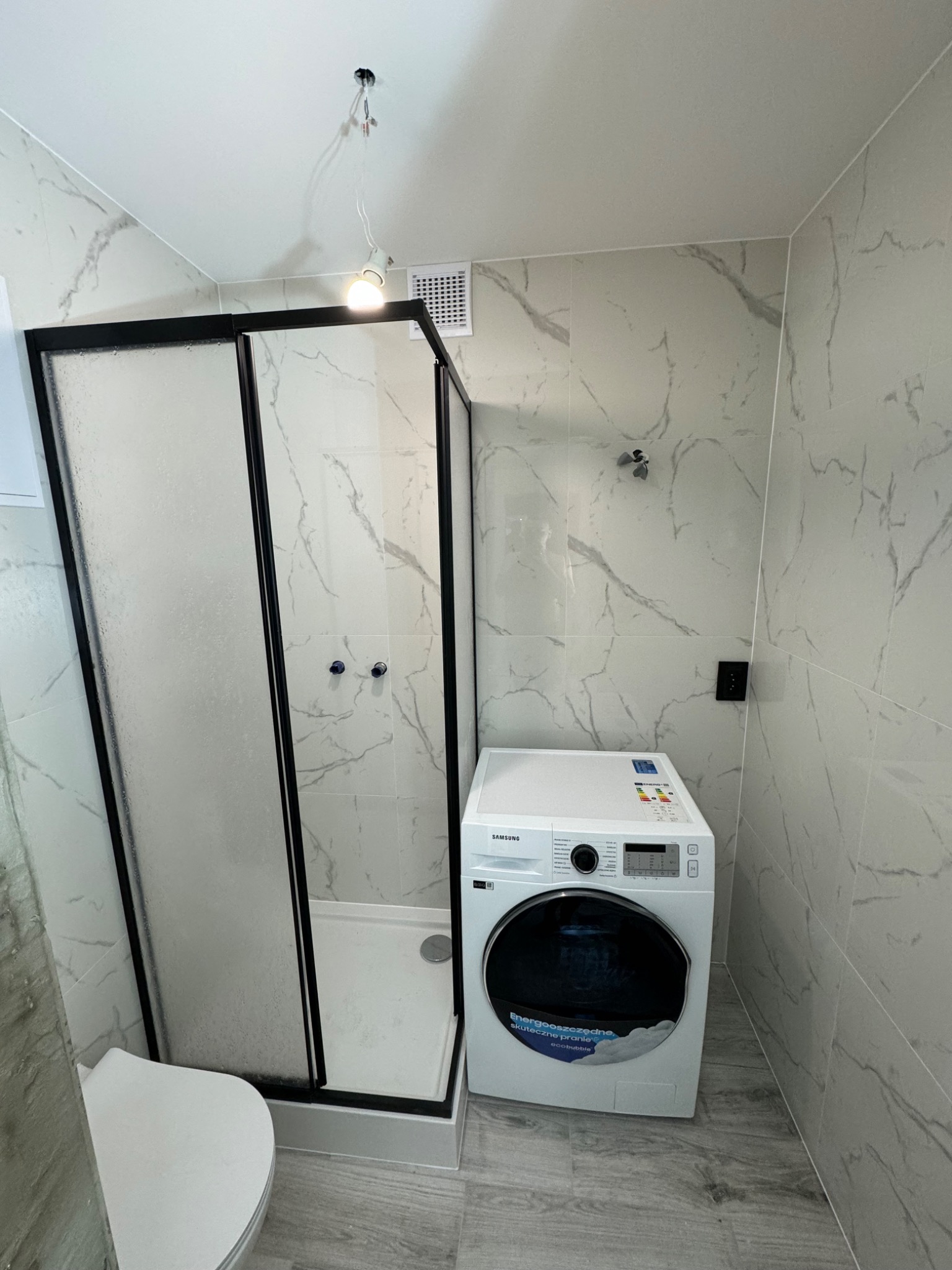 Nowoczesna łazienka z kabiną prysznicową z czarną ramą, białą pralką Samsung i płytkami imitującymi marmur na ścianach i podłodze.