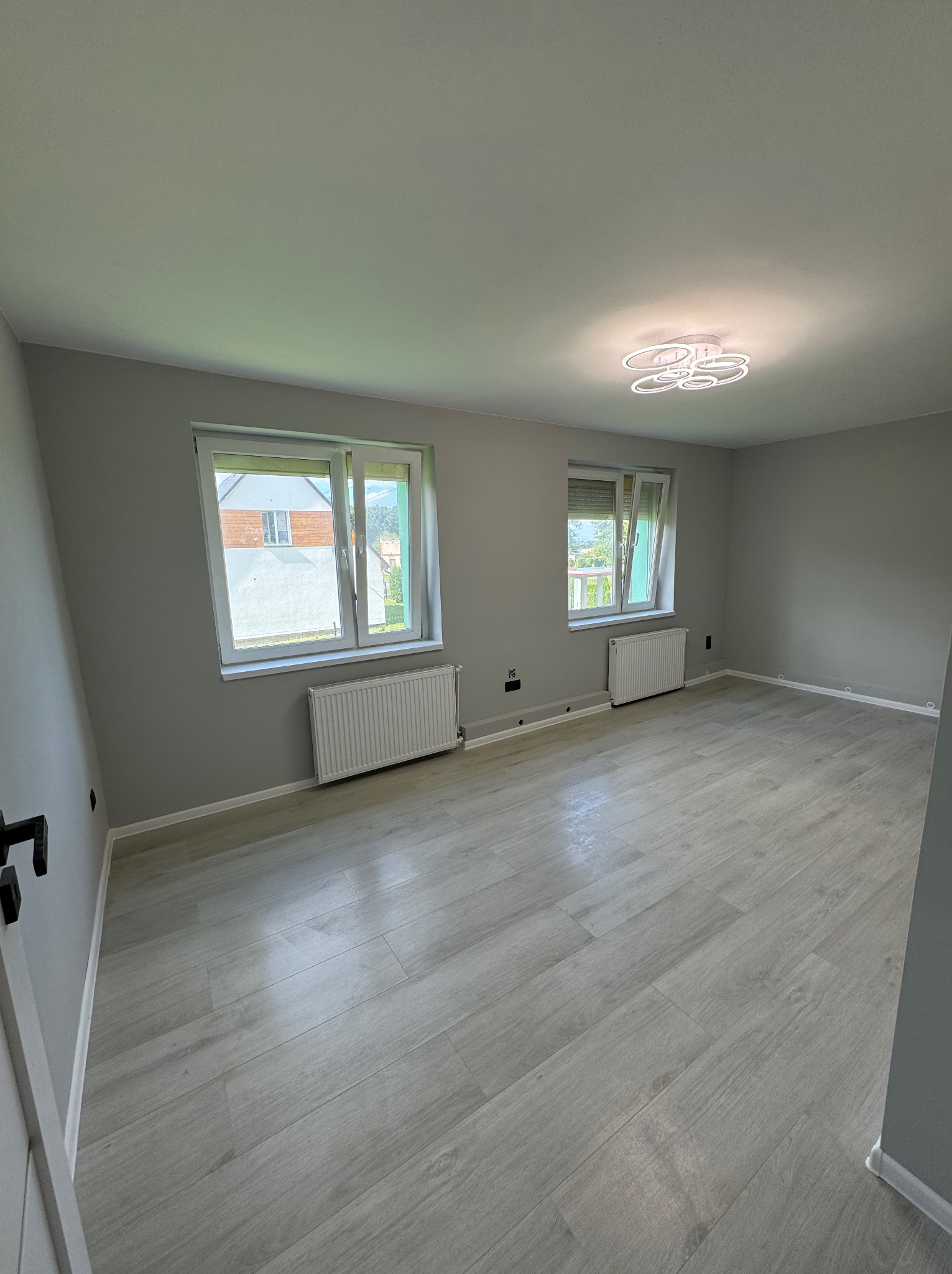 Pomalowane na szaro wnętrze pokoju z dwiema oknami, białymi grzejnikami i panelami podłogowymi w odcieniach szarości oraz nowoczesną lampą sufitową.