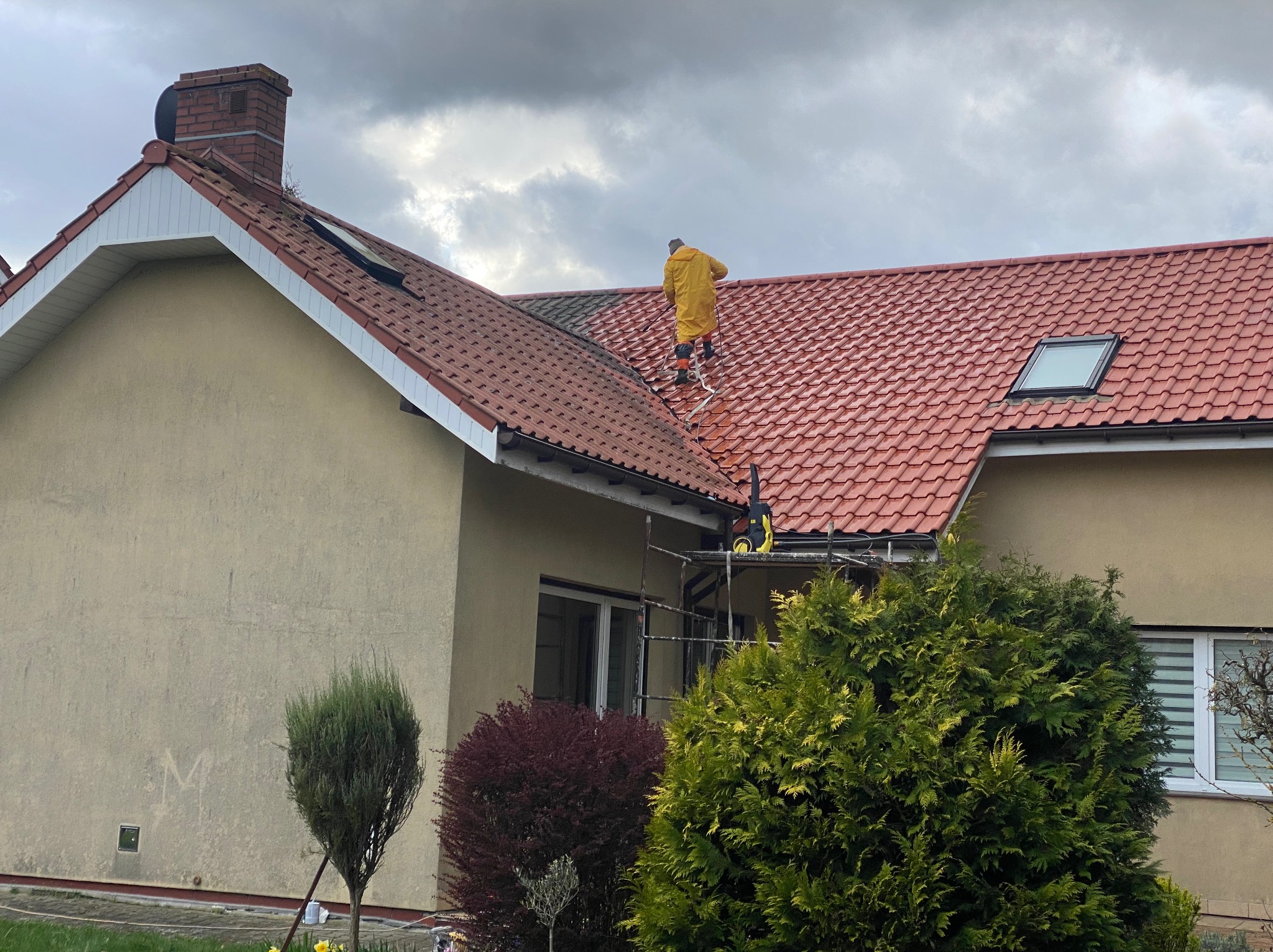 Pracownik w żółtym kombinezonie czyści czerwoną dachówkę domu jednorodzinnego za pomocą myjki ciśnieniowej, stojąc na drabinie linowej na tle pochmurnego nieba.