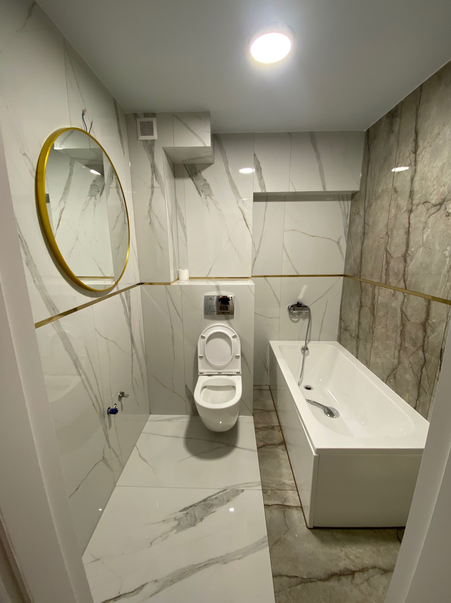 Nowoczesna łazienka z marmurowymi płytkami, złotymi akcentami, okrągłym lustrem i białą armaturą: toaleta i prostokątna wanna.