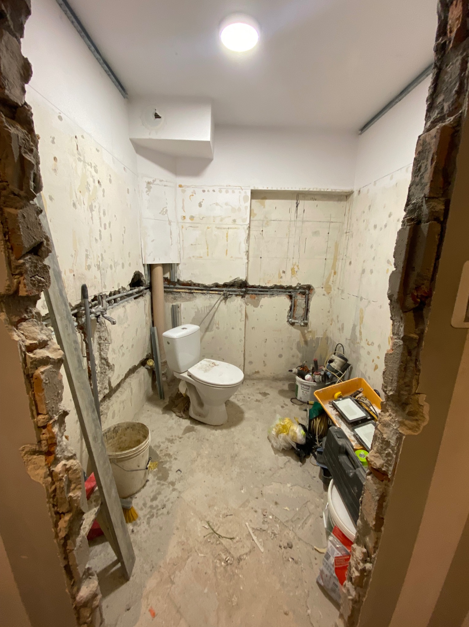 Łazienka w trakcie remontu: obnażone rury, uszkodzone ściany z resztkami płytek, stojąca toaleta, widoczne narzędzia i wiadro, częściowo wyburzone ściany przy wejściu.