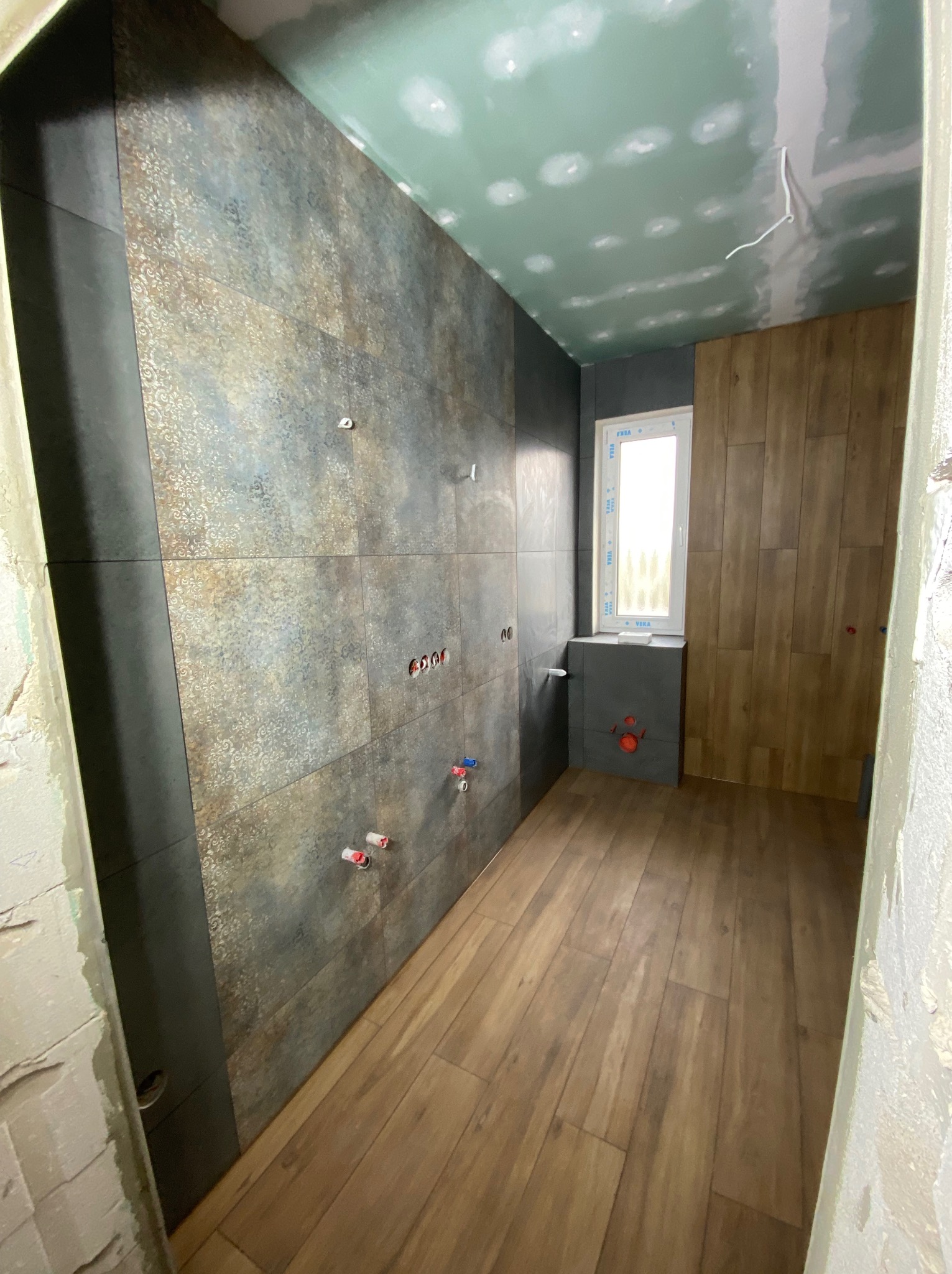 Łazienka w trakcie remontu: położone płytki imitujące metal z ornamentami na ścianie, drewnopodobne panele na podłodze, sufit z widocznymi śladami po szpachlowaniu, wystające rury instalacji wodnej.