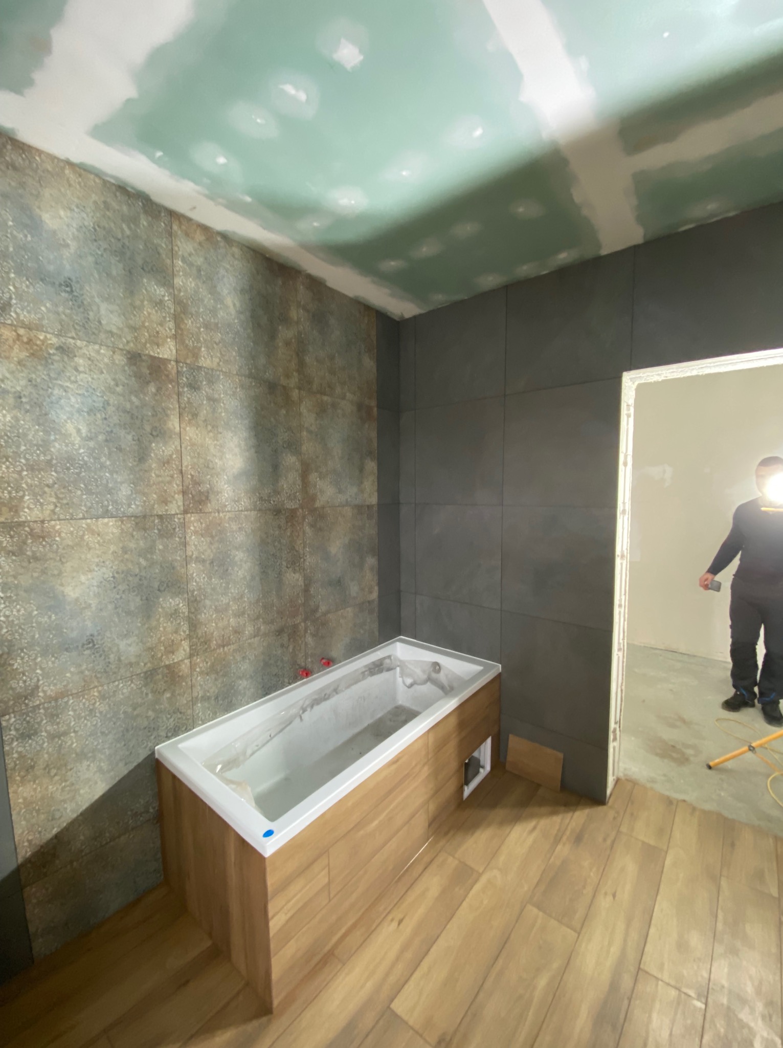 Wnętrze łazienki w trakcie remontu: położona glazura w odcieniach szarości i brązu, obudowana wanna, sufit z płyt gipsowo-kartonowych przed szpachlowaniem, podłoga wyłożona panelami imitującymi...