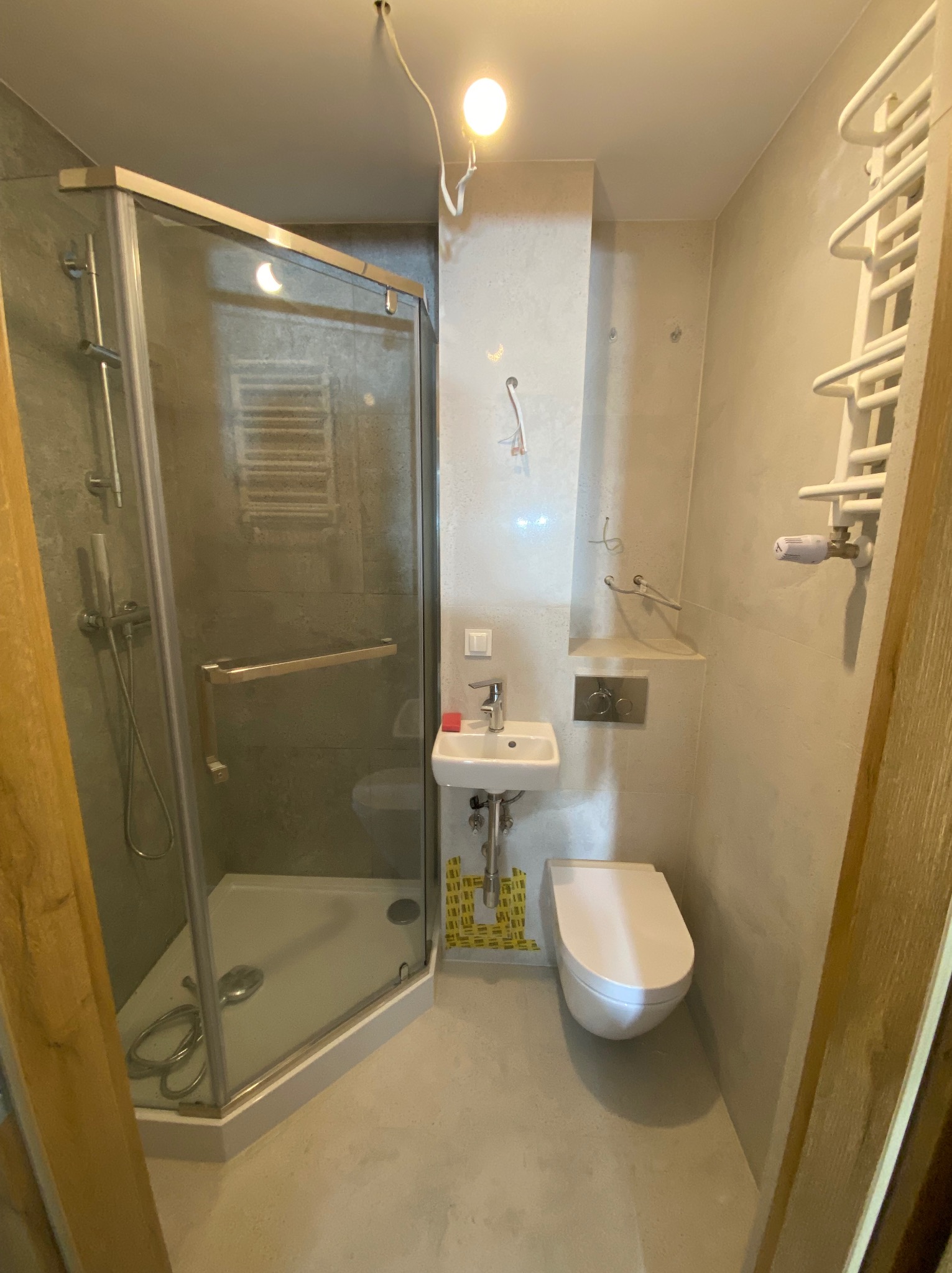 Nowoczesna łazienka z kabiną prysznicową, wiszącą toaletą i małą umywalką, wykończona w szarościach z akcentem żółtych płytek dekoracyjnych.