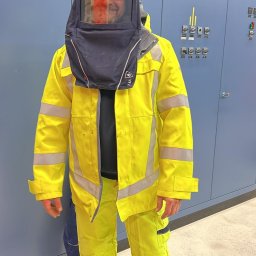 WOMAX Wojciech Wojs - Elektryk w stroju ochronnym, z osłoną na twarzy, stojący przed szafą sterowniczą w pomieszczeniu technicznym.