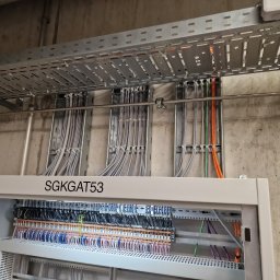 WOMAX Wojciech Wojs - Rozdzielnica elektryczna z licznymi przewodami wewnątrz, ułożonymi w rzędach i oznaczonymi kolorami, nad nią koryto kablowe z przewodami przytwierdzonymi opaskami zaciskowymi, na tle surowej ściany...