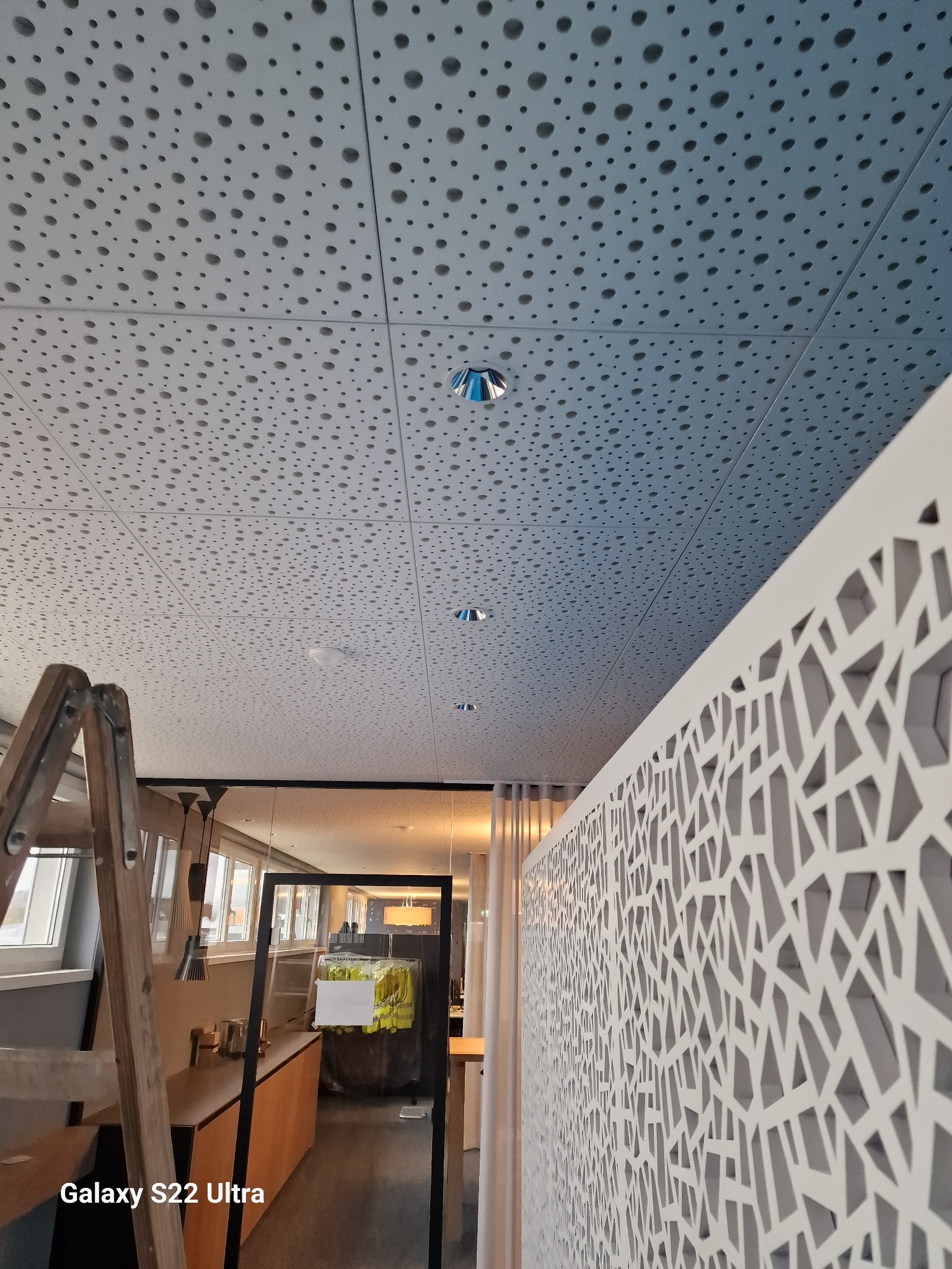 Panelowy sufit akustyczny z perforacją, widoczne oświetlenie, drabina i dekoracyjna ścianka działowa z ażurowym wzorem.