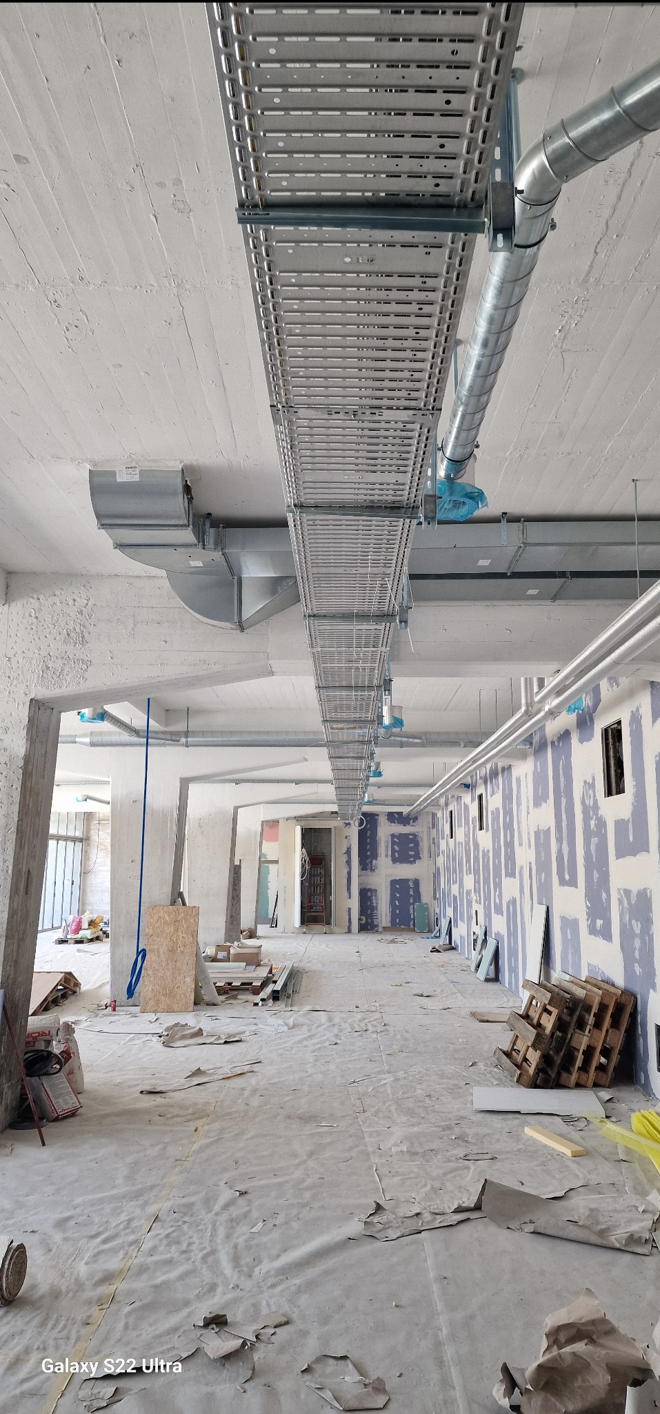 Wnętrze w trakcie remontu z widocznymi metalowymi korytkami kablowymi pod sufitem, instalacje wentylacyjne, ściany przygotowane do malowania i zabezpieczona podłoga.