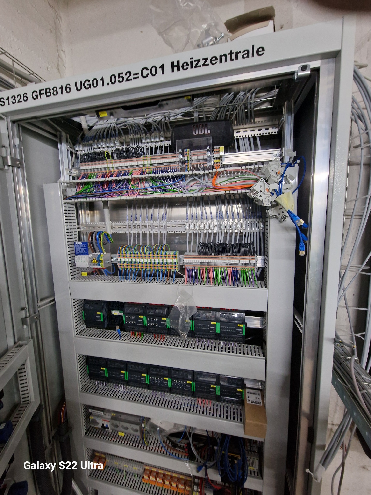 Rozdzielnica elektryczna z widocznymi przewodami, sterownikami PLC i elementami zabezpieczeń, oznaczona napisem 'Heizzentrale' na górze.