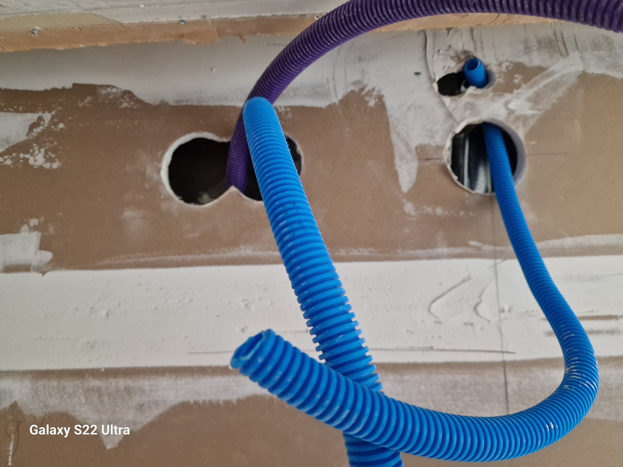 Instalacja elektryczna w trakcie prac wykończeniowych: niebieskie i fioletowe peszle wychodzące z otworów w ścianie z płyt kartonowo-gipsowych, widoczne ślady szpachlowania.