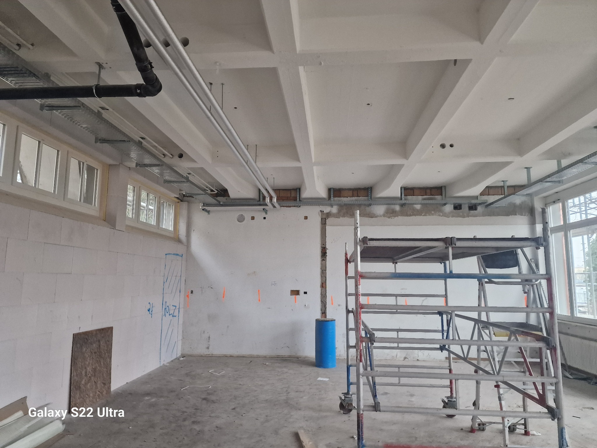 Wnętrze w trakcie remontu: surowy betonowy sufit z widocznymi instalacjami, ściany z bloków z zaznaczeniami, rusztowanie, niebieska beczka i widoczne okablowanie elektryczne w korytach.