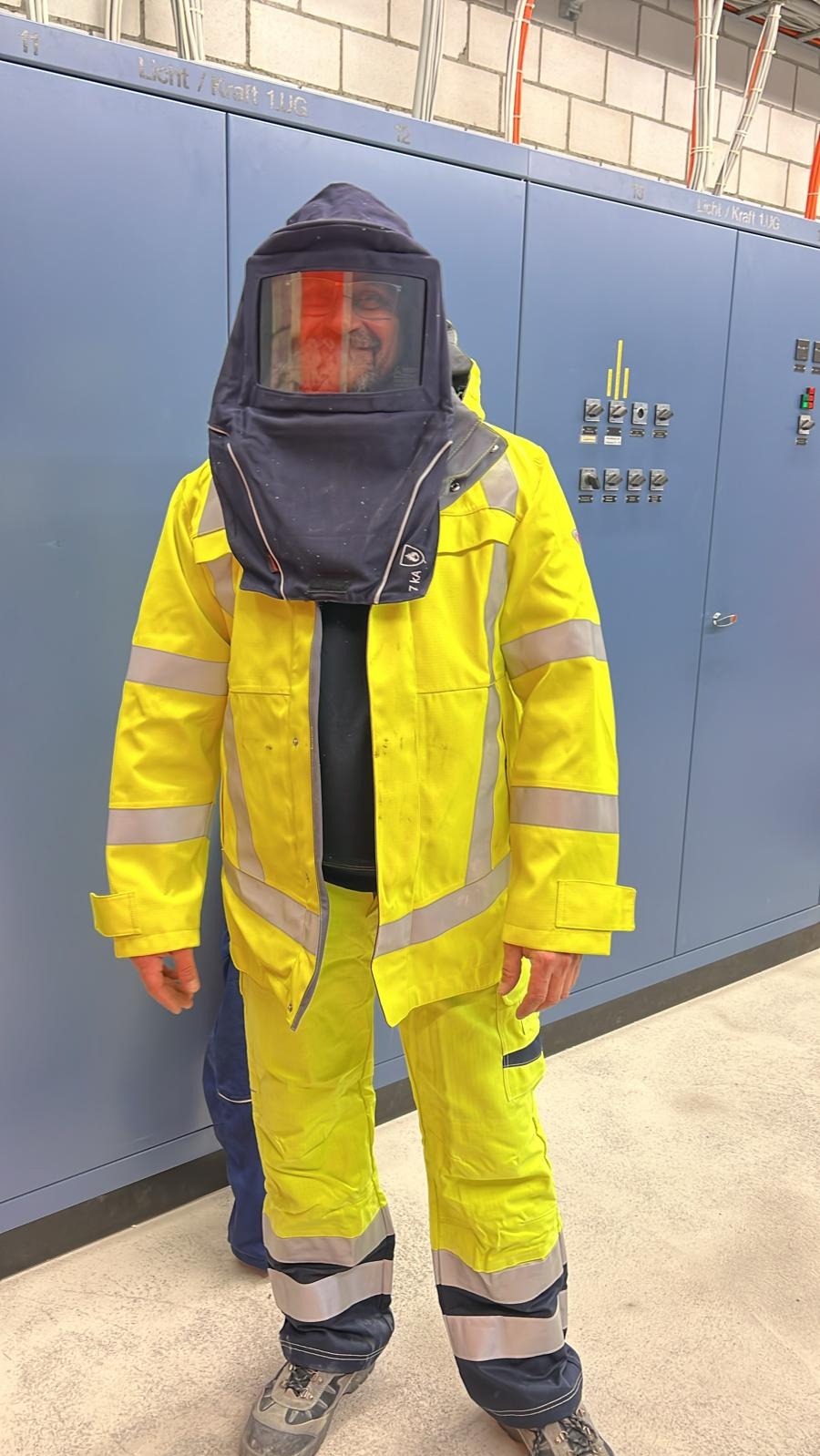 Elektryk w stroju ochronnym, z osłoną na twarzy, stojący przed szafą sterowniczą w pomieszczeniu technicznym.