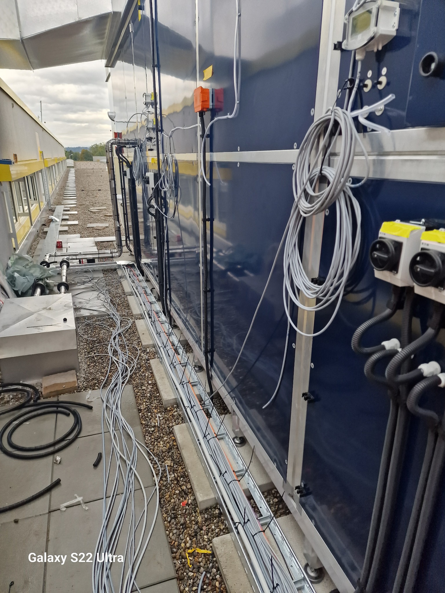 Montaż przewodów elektrycznych na dachu budynku, widoczne kable w korytach, zwisające przewody i urządzenia sterujące na bocznej ścianie.