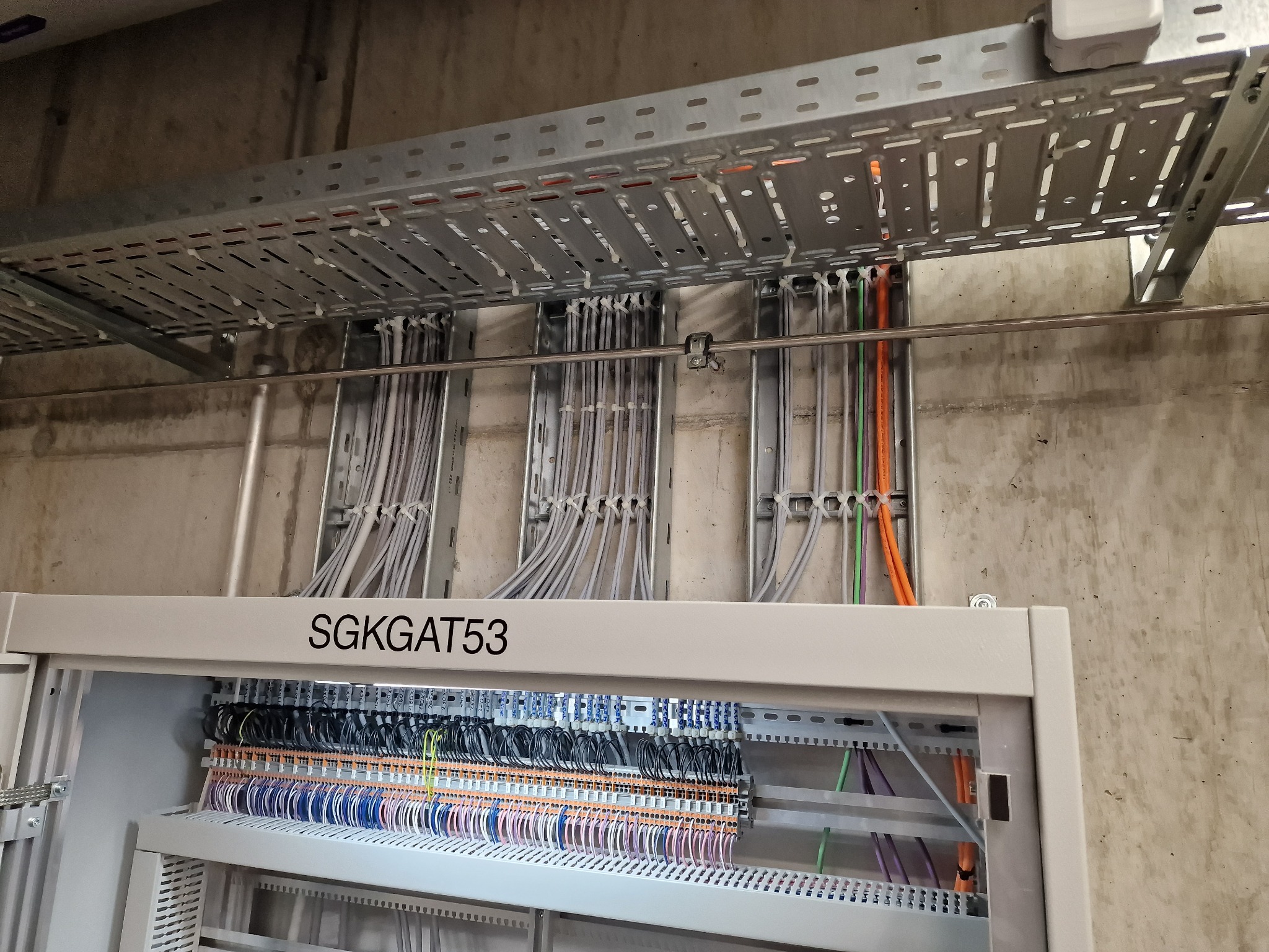 Rozdzielnica elektryczna z licznymi przewodami wewnątrz, ułożonymi w rzędach i oznaczonymi kolorami, nad nią koryto kablowe z przewodami przytwierdzonymi opaskami zaciskowymi, na tle surowej ściany...