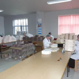 Pracownicy w białych fartuchach i czepkach ręcznie układają czekoladki w pudełka w jasnym pomieszczeniu produkcyjnym; widoczne stosy zapakowanych produktów i puste opakowania.