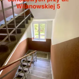 Klatka schodowa po remoncie: ściany pomalowane na biało i brązowo, schody szare, okno z widokiem na zieleń, metalowa balustrada. Perspektywa z góry.