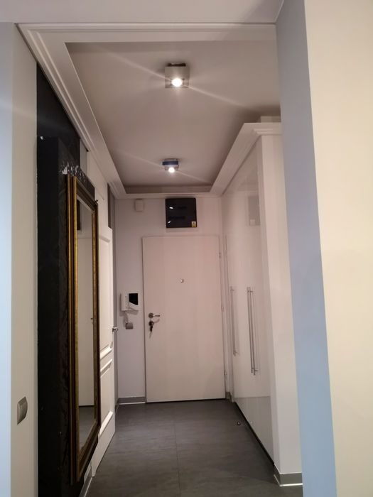 Korytarz po remoncie z białymi ścianami, szarą podłogą, lustrem w złotej ramie i ozdobnym oświetleniem sufitowym.