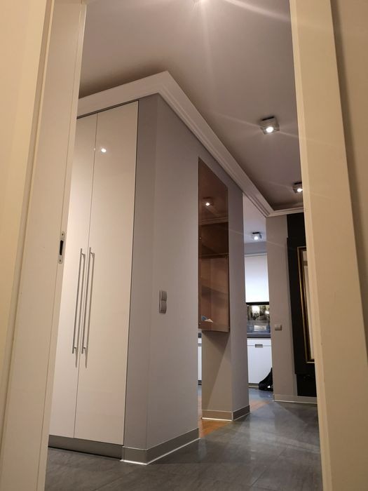 Nowoczesne wnętrze przedpokoju z szafą w zabudowie, szarą ścianą z listwą przypodłogową z oświetleniem LED i widokiem na kuchnię w tle.