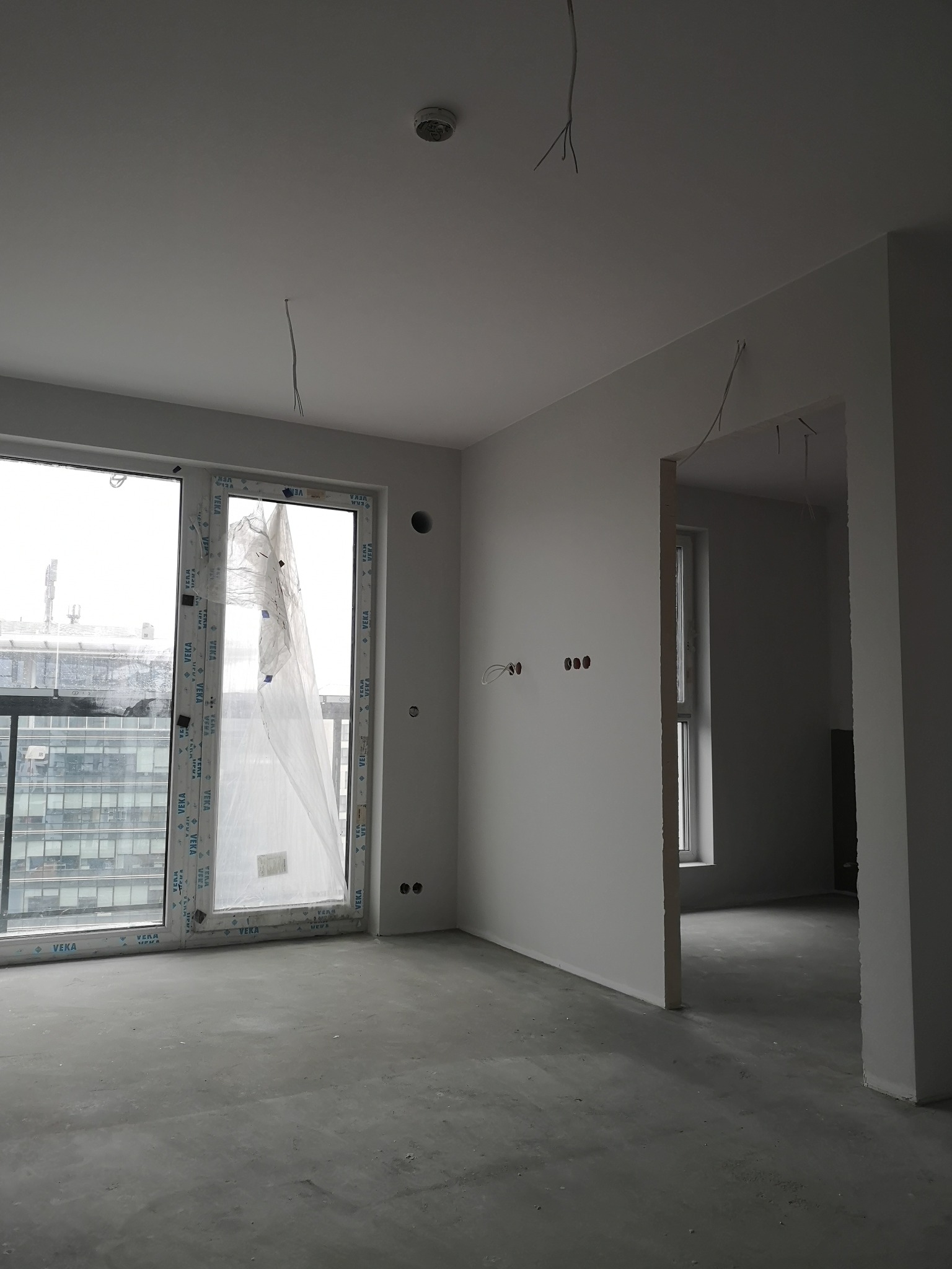Surowe wnętrze mieszkania w stanie deweloperskim po nałożeniu gładzi, widoczne nieosłonięte przewody elektryczne, okno z częściowo zdjętą folią ochronną, surowa wylewka podłogowa.