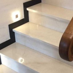 Komi - Schody wewnętrzne wykończone jasnym marmurem z czarnymi akcentami i drewnianą poręczą, oświetlone punktowo.