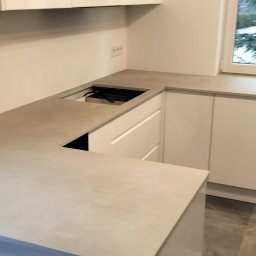 Komi - Nowoczesna, minimalistyczna kuchnia z białymi szafkami i beżowym blatem, widoczne gniazdka elektryczne na ścianie, fragment okna z widokiem na zieleń i poziomica na blacie.