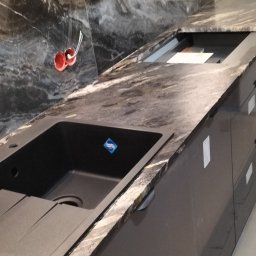 Komi - Ciemny, granitowy blat kuchenny z wbudowanym czarnym zlewozmywakiem, pod marmurową ścianą i drewnianymi szafkami. Widoczne wystające kable elektryczne.