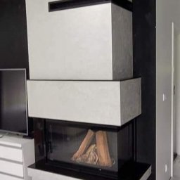 Komi - Nowoczesny kominek narożny z obudową w odcieniach szarości i czerni, z widocznym paleniskiem i drewnem wewnątrz, na tle marmurowej podłogi i czarnej ściany.