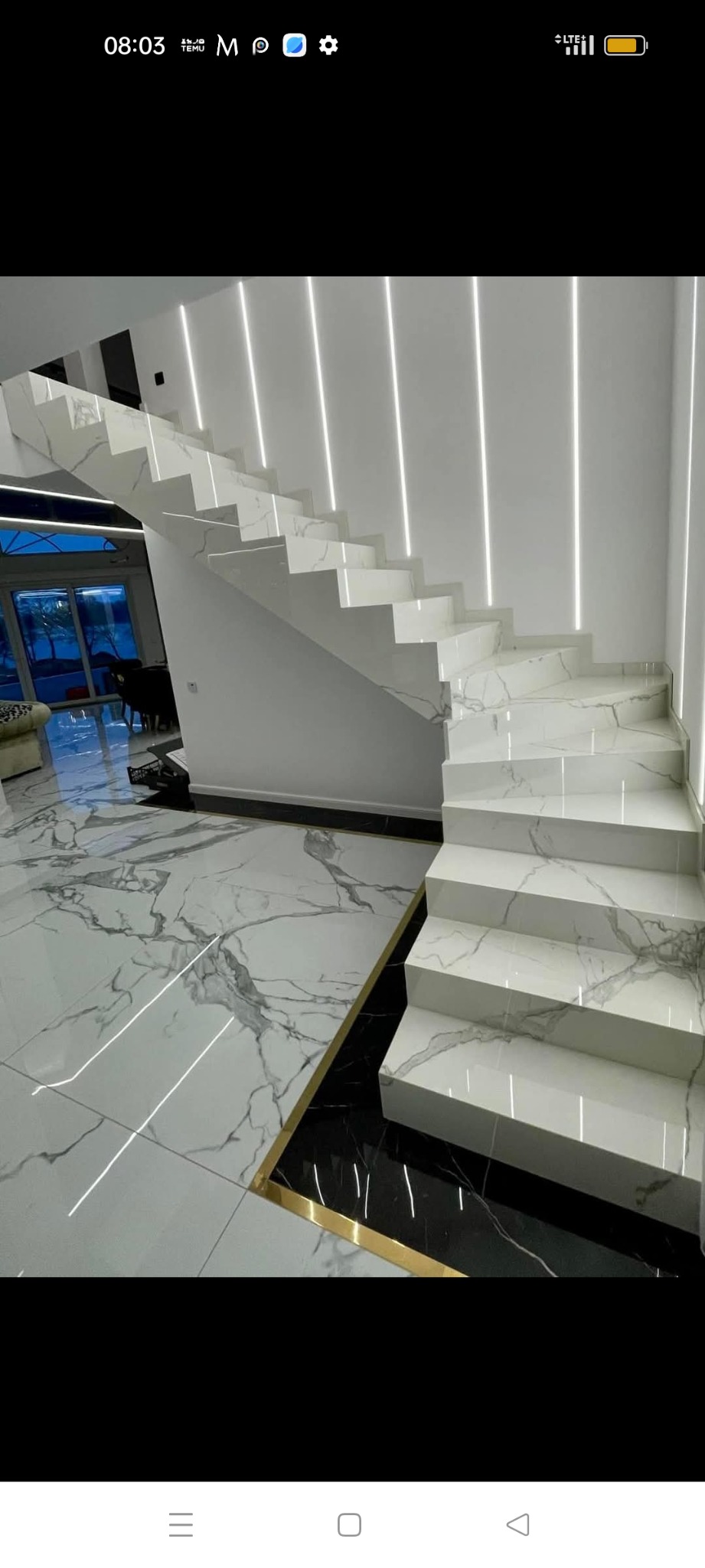 Schody wewnętrzne wykończone białym marmurem z szarymi żyłami, podświetlone pionowymi pasami LED, z podłogą z płytek imitujących marmur i czarną wstawką z obramowaniem w kolorze złotym u podstawy...