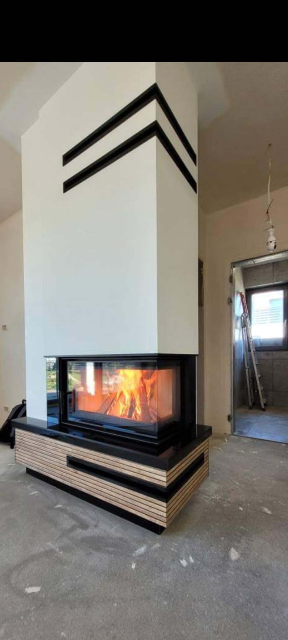 Nowoczesny kominek narożny z paleniskiem, obudowany płytami z jasnego drewna i czarnymi elementami, w surowym wnętrzu z widoczną drabiną i niedokończonym sufitem.