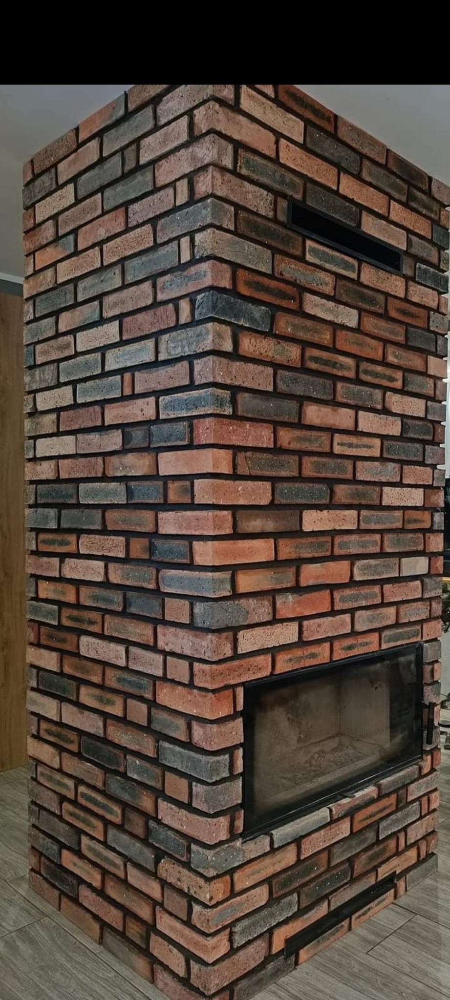 Murowany kominek z cegły klinkierowej o zróżnicowanej kolorystyce, z czarną ramą paleniska i kratką wentylacyjną u góry.
