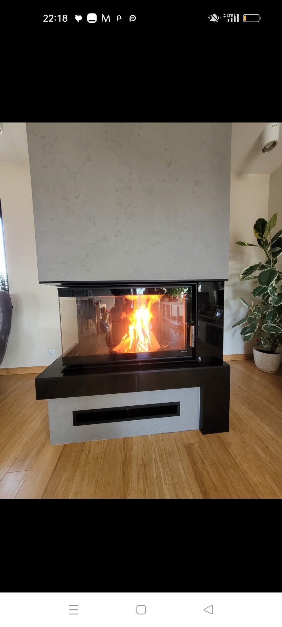 Nowoczesny kominek narożny z szybą, obudowany płytami w odcieniach szarości i czerni, z płonącym wewnątrz ogniem, na tle parkietu i rośliny doniczkowej.