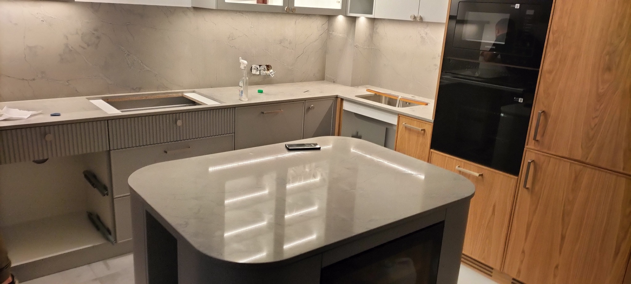 Montaż szarej kuchni z wyspą i marmurowym blatem, widoczne niedokończone podłączenia elektryczne i hydrauliczne.