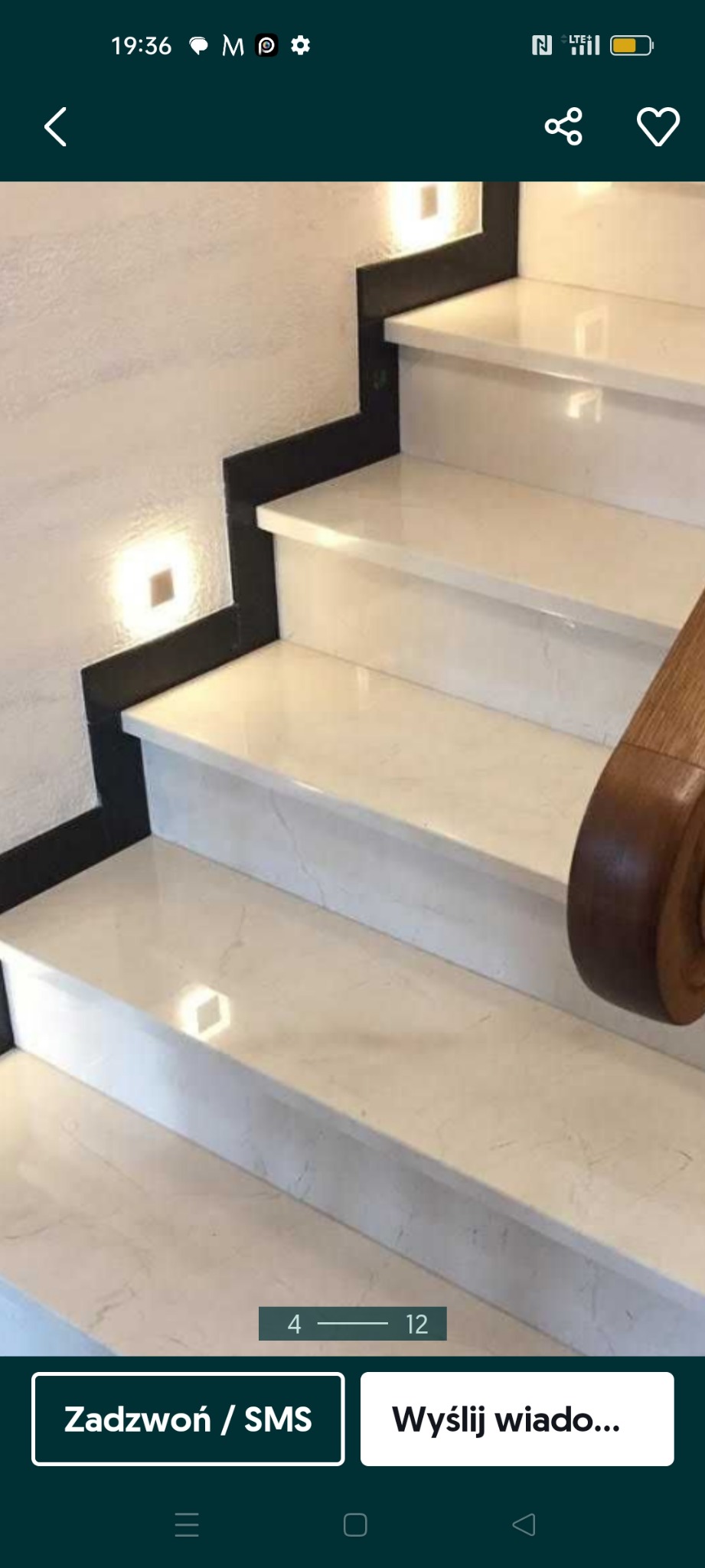 Schody wewnętrzne wykończone jasnym marmurem z czarnymi akcentami i drewnianą poręczą, oświetlone punktowo.