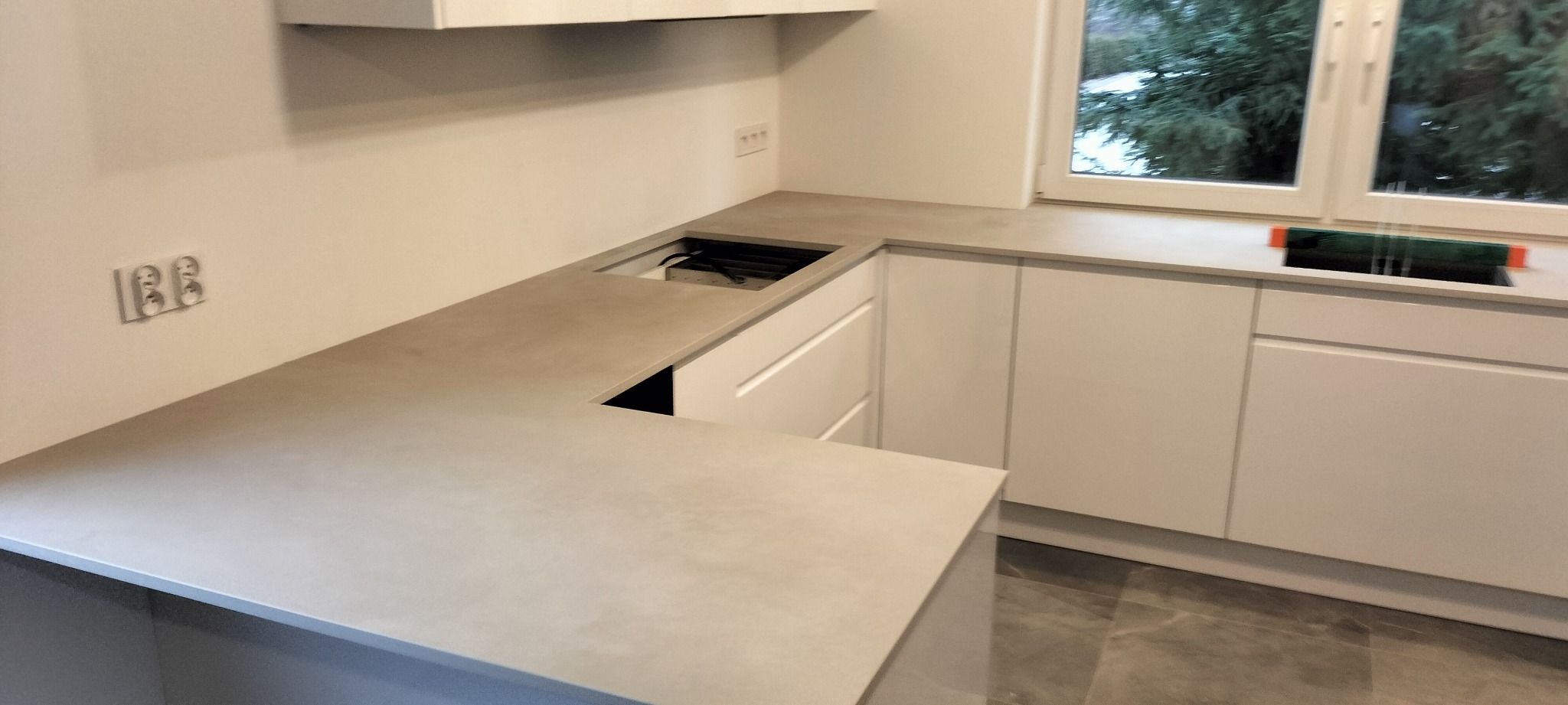 Nowoczesna, minimalistyczna kuchnia z białymi szafkami i beżowym blatem, widoczne gniazdka elektryczne na ścianie, fragment okna z widokiem na zieleń i poziomica na blacie.