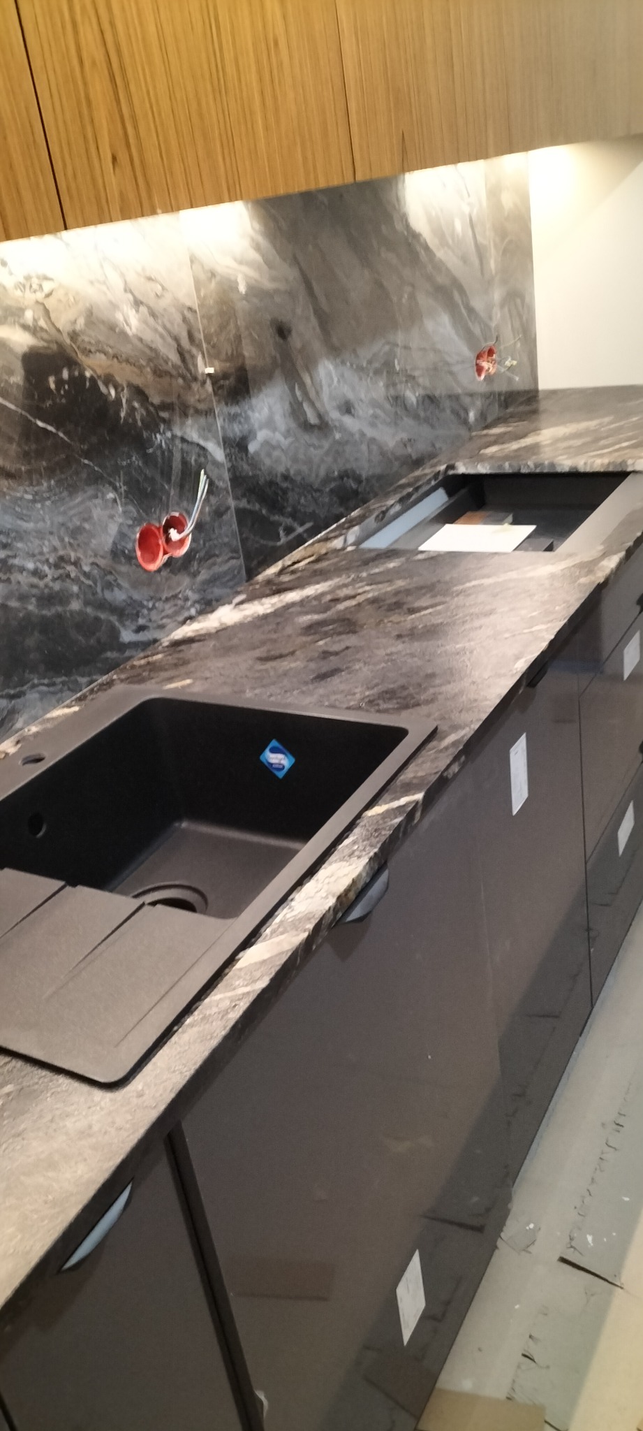 Ciemny, granitowy blat kuchenny z wbudowanym czarnym zlewozmywakiem, pod marmurową ścianą i drewnianymi szafkami. Widoczne wystające kable elektryczne.