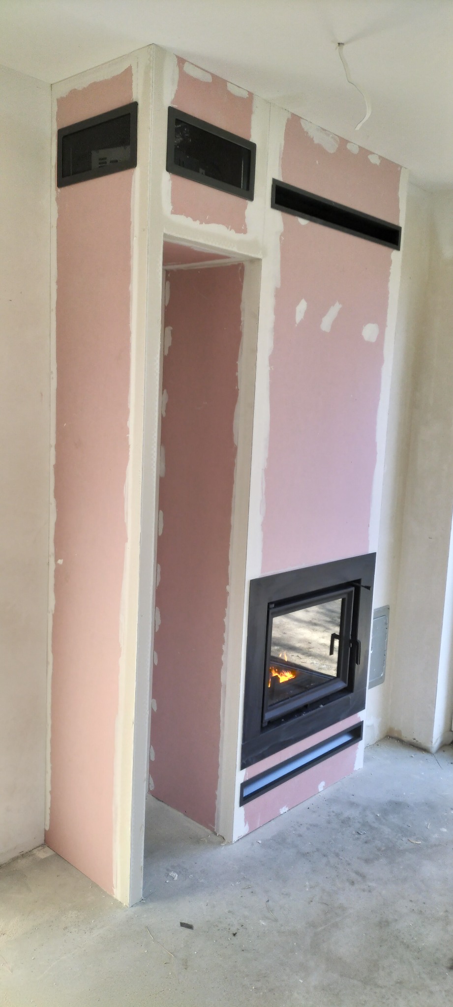 Wnętrze w trakcie wykończenia z zabudowanym kominkiem z czarnym wkładem, obudowa z płyt karton-gips, widoczne kratki wentylacyjne.