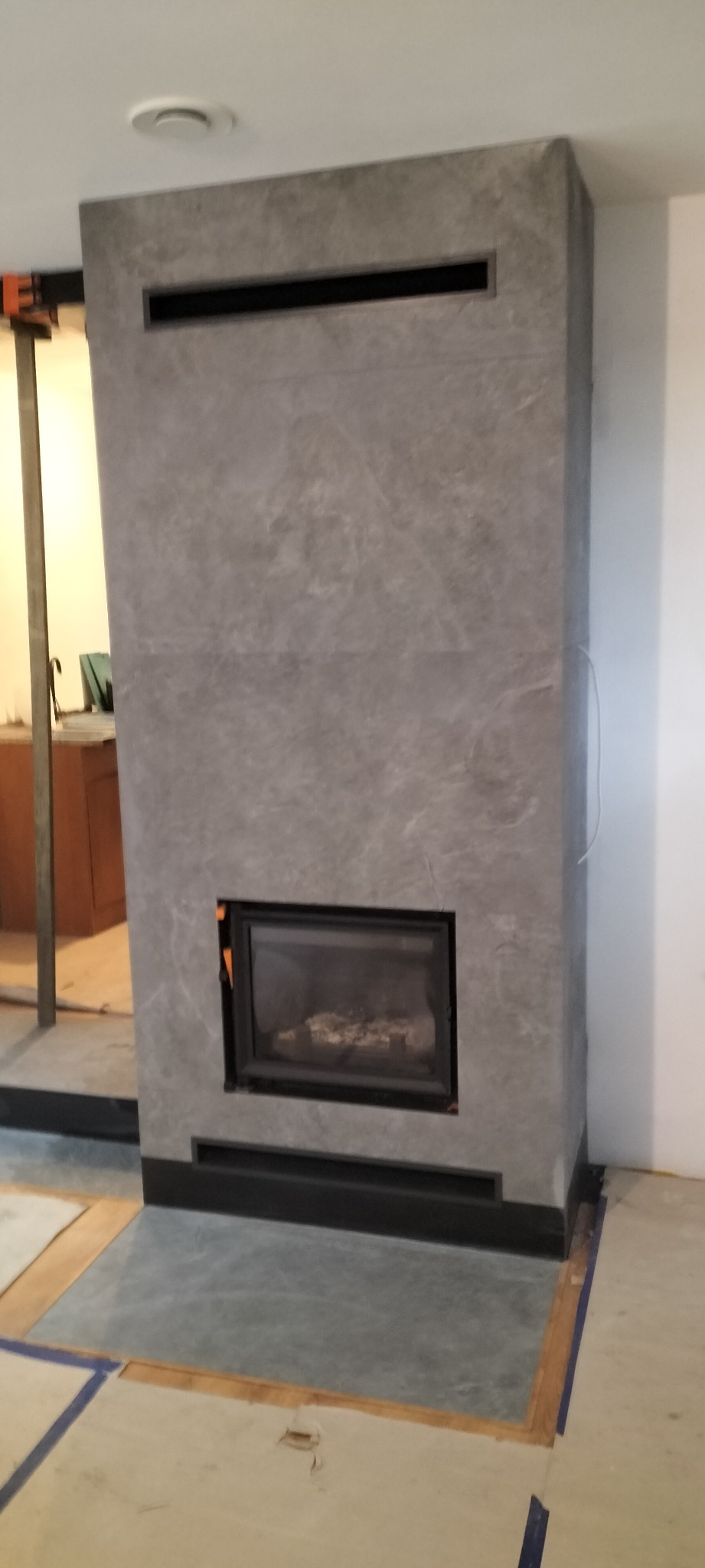 Szary kominek obudowany płytami imitującymi kamień, z czarną ramą paleniska i poziomymi szczelinami wentylacyjnymi, w trakcie montażu, widoczne elementy konstrukcyjne i zabezpieczenia podłogi.