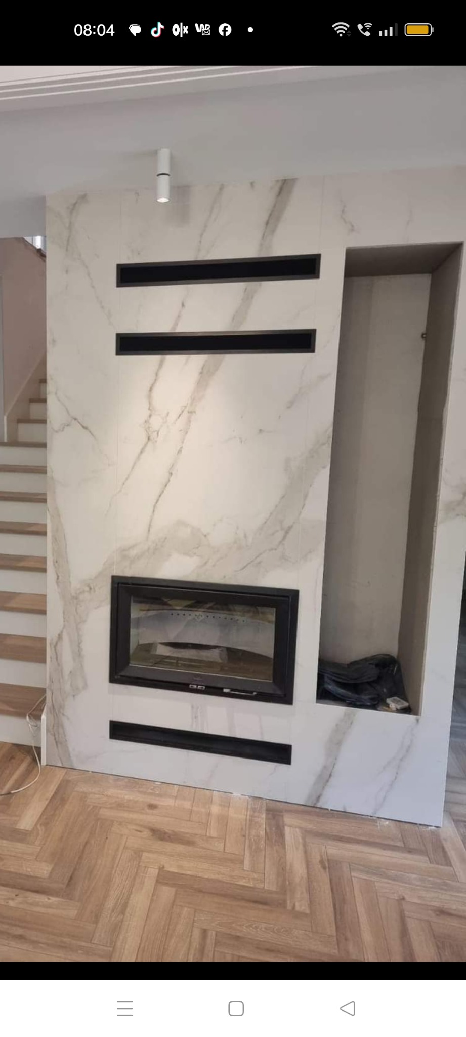 Nowoczesny kominek z czarną obudową, wbudowany w ścianę z marmurowym wzorem, z dodatkowymi czarnymi elementami dekoracyjnymi i wnęką po prawej stronie, parkiet w jodełkę na podłodze.